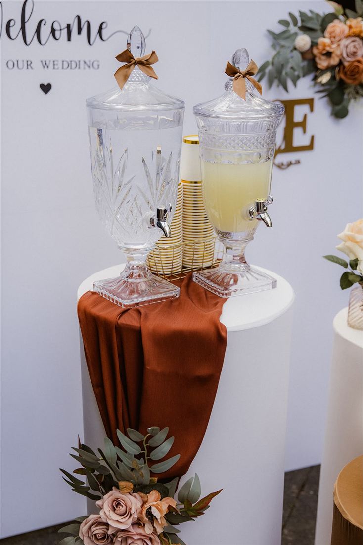 Getränkestation einer Hochzeit: Kristallspender mit Limonade auf weißer Säule, Tuch in Terracotta und Rosen