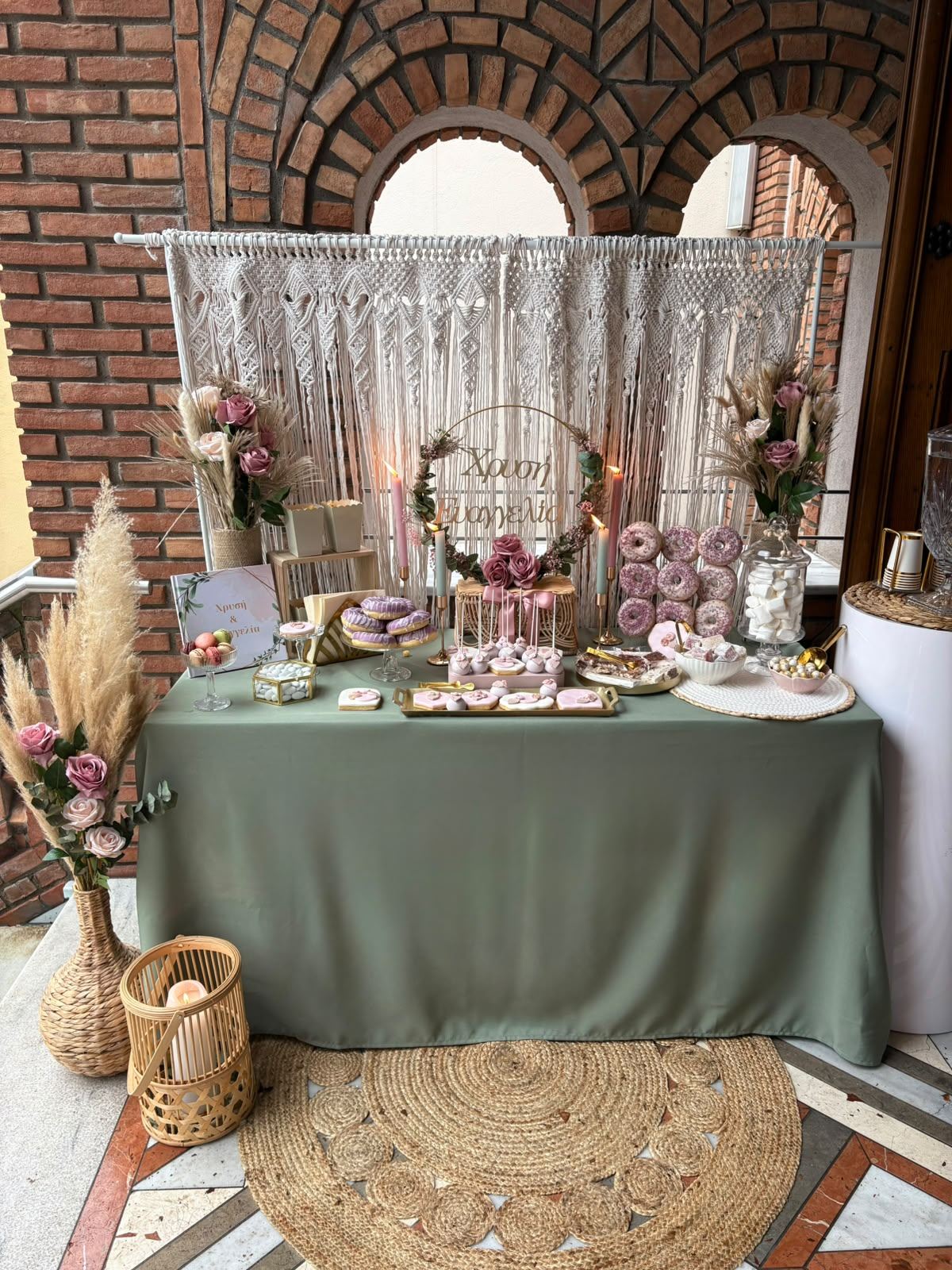 Sweet Table mit Makramee-Rückwand, salbeigrüner Tischdecke, rosa Rosen und Donut-Wand vor Backstein-Rundbögen